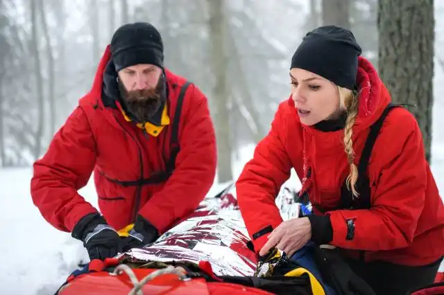 Paramedics from mountain rescue service provide operation outdoors in winter in forest, injured person in stretcher.