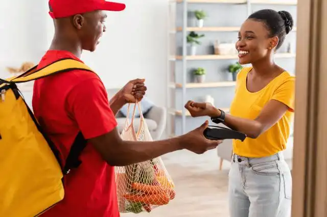 African American Woman Paying Supermarket Delivery Guy Using Smartwatch At Home. Female Customer Putting Watch To Payment Ter