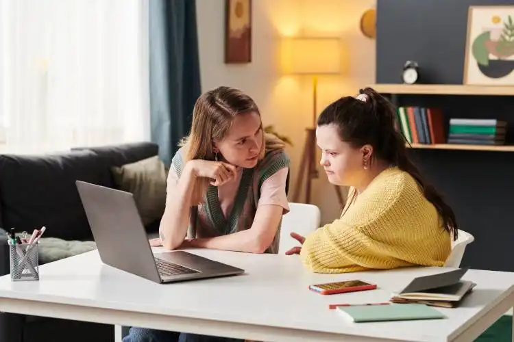 Young woman with Down syndrome and her university friend sitting at table working together on project looking at laptop scree