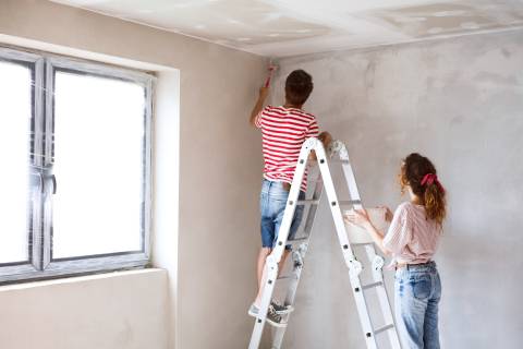 Beautiful young couple standing on ladder painting walls in their new house using paint brushes. Home makeover and renovation