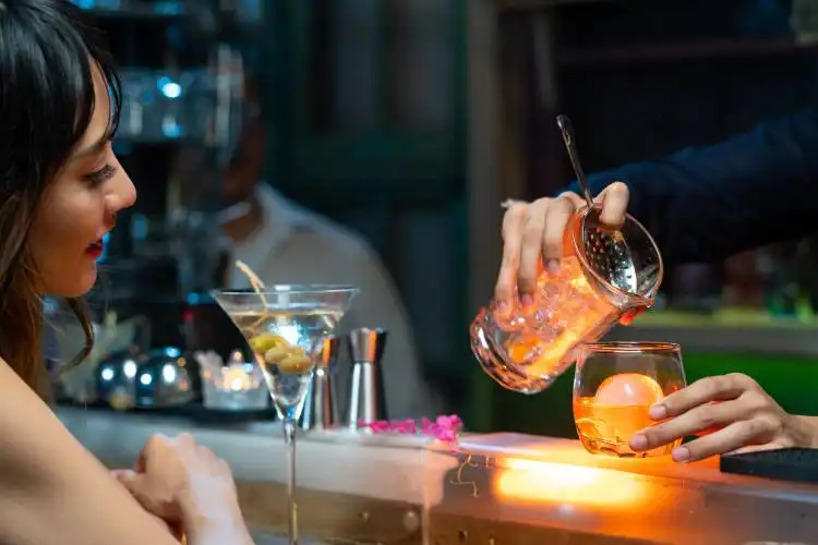 Confidence Asian woman sitting at bar counter drinking alcoholic drink and talking to barman in nightclub. Male mixologist ba