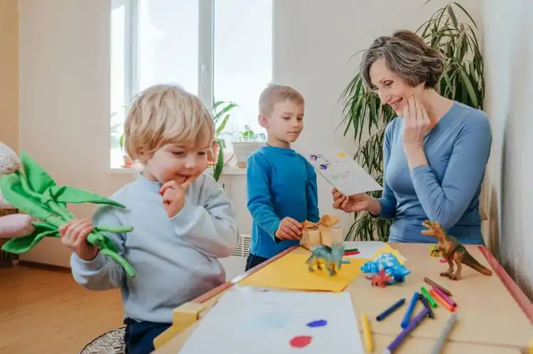 Grandchildren presenting to their grandmother a handmade drawing postcard and flowers indoor. Selective focus.