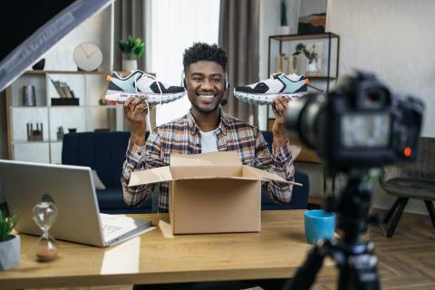 Smiling african man sitting at table with new sneakers in hands and recording video review on professional camera. Concept of