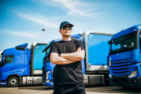 Professional truck driver with hat and sunglasses confidently standing in front of big and modern truck. Bright sunny day. Pe