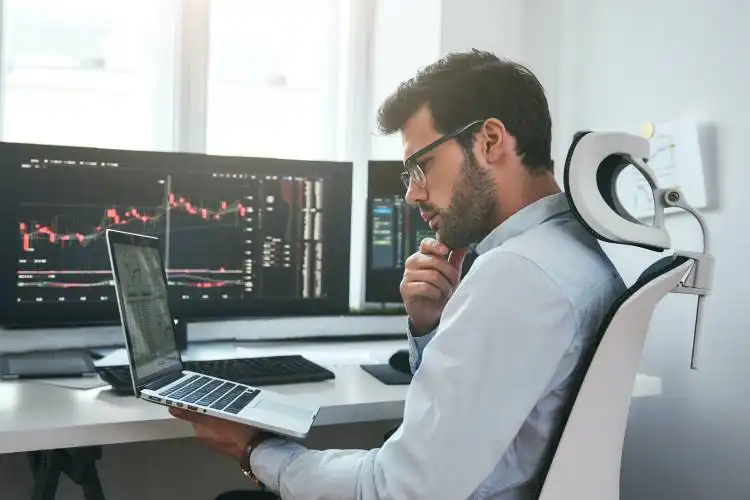 Busy day. Professional male trader wearing eyeglasses analyzing financial market via laptop while sitting in front of compute